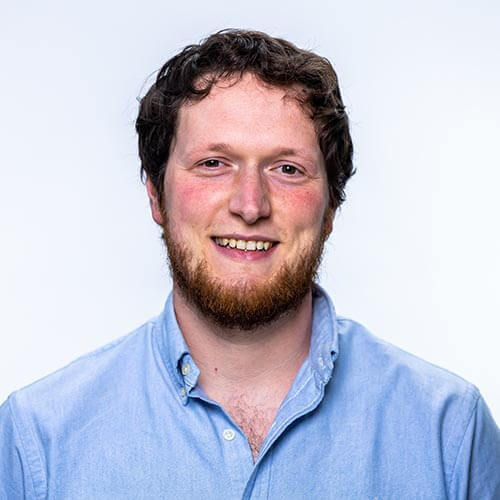 A man with short brown hair and a brown beard wearing a blue shirt against a plain backdrop