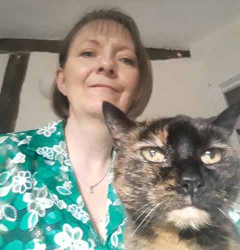 short-haired brunette woman wearing green flowery dress standing behind tortoiseshell cat