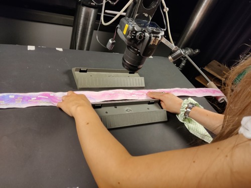 woman holding long, thin piece of paper on table and filming it with camera from above